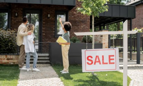 african american real estate agent showing house to hugging couple near for sale signboard