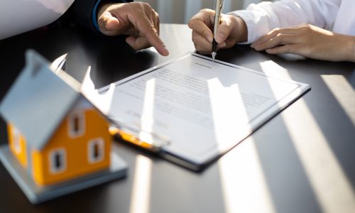 Businessman in suit in his office showing home insurance policy and pointing with a pen where the policyholder must to sign. Insurance agent consulting real estate insurance detail to customer.