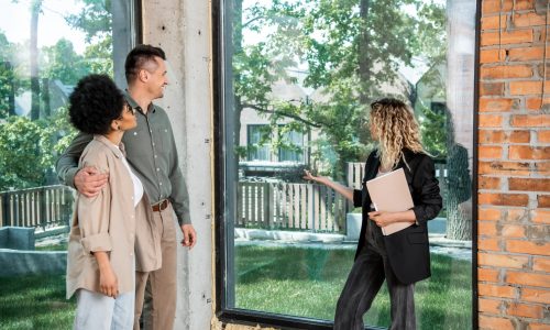 real estate agent with folder pointing with hand at window in new cottage near interracial couple